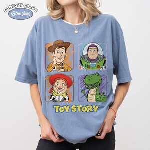 May include: Blue short-sleeved t-shirt featuring four character portraits from Toy Story: Woody, Buzz Lightyear, Jessie, and Rex. The words "TOY STORY" are printed in blue below the characters. The shirt has the words "Comfort Color" and "Blue Jean" printed on the top left.