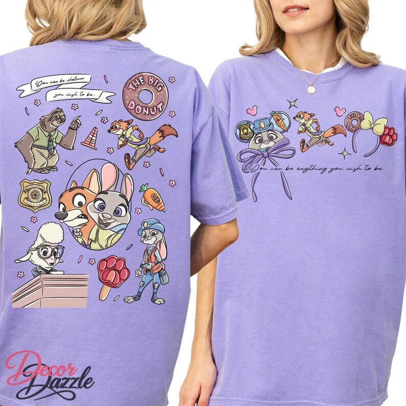 May include: A light purple t-shirt featuring cartoon characters and the text "You can be whatever you wish to be." The design includes a donut, a police badge, and various animal characters. The shirt has a relaxed fit.