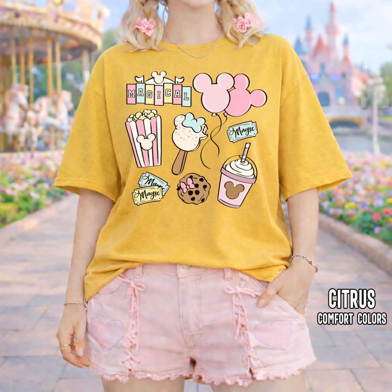 May include: Mustard yellow t-shirt with a graphic design featuring the word "MAGICAL" and cartoon illustrations of popcorn, ice cream, balloons, a cookie, and a drink. The shirt is paired with pink shorts. The text "CITRUS COMFORT COLORS" is in the bottom right corner.