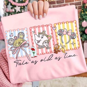 Puede incluir: Camiseta rosa claro con ilustraciones de personajes de La Bella y la Bestia, incluyendo a la Sra. Potts, Chip y una rosa. La camiseta tiene la frase "Tale as old as time" en escritura negra.