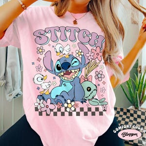 May include: Pink t-shirt featuring the word "Stitch" in a retro font above a cartoon image of Stitch, two ducks, and a plush toy. The design includes flowers and a checkered pattern at the bottom.