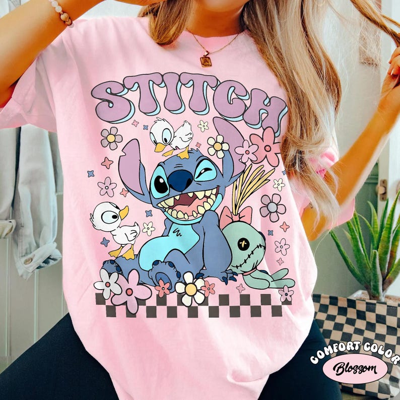 May include: Pink t-shirt featuring the word "Stitch" in a retro font above a cartoon image of Stitch, two ducks, and a plush toy. The design includes flowers and a checkered pattern at the bottom.