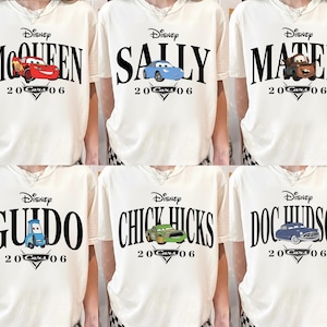 Op de afbeelding: Cr&egrave;mekleurige t-shirts met Disney Cars-personages. Elk shirt toont de naam van een ander personage in grote zwarte letters, samen met een bijbehorende autografiek en de tekst "20 Cars 06".