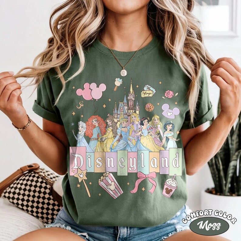 May include: Green t-shirt with a Disneyland graphic featuring Disney princesses, a castle, and themed icons. The word "Disneyland" is in pastel colours. The shirt is labelled "Comfort Color Moss".