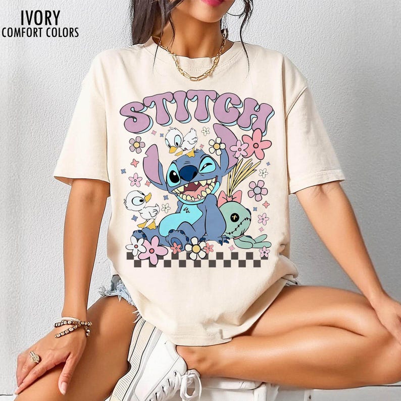 May include: Ivory-colored t-shirt featuring a graphic of the character Stitch with the word "STITCH" in a purple bubble font. The design includes flowers, ducks, and a green plush toy. The shirt is labeled "IVORY COMFORT COLORS".