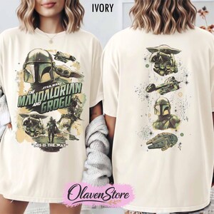 May include: Ivory-colored t-shirt featuring a graphic design from the Star Wars series, "The Mandalorian and Grogu." The front displays characters and text, while the back showcases additional imagery. The shirt has a relaxed fit.