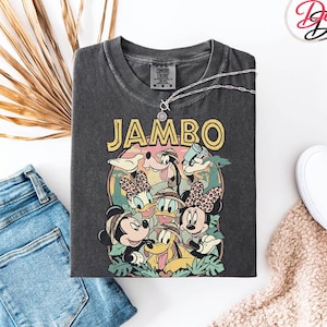 Puede incluir: Camiseta gris oscuro con un gr&aacute;fico colorido de personajes de Disney, incluyendo Mickey, Minnie, Donald y Goofy, con la palabra "JAMBO" en amarillo. La camiseta se combina con vaqueros azules, zapatos blancos y un collar plateado.