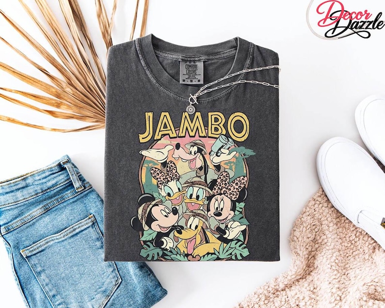 Puede incluir: Camiseta gris oscuro con un gr&aacute;fico colorido de personajes de Disney, incluyendo Mickey, Minnie, Donald y Goofy, con la palabra "JAMBO" en amarillo. La camiseta se combina con vaqueros azules, zapatos blancos y un collar plateado.