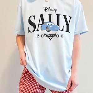 Op de afbeelding: Lichtblauw T-shirt met het Disney Cars-personage Sally. Het shirt heeft het Disney-logo boven de naam "SALLY" in grote zwarte letters, met "2006" en een autografiek eronder. De persoon draagt een rood-wit geruite short.
