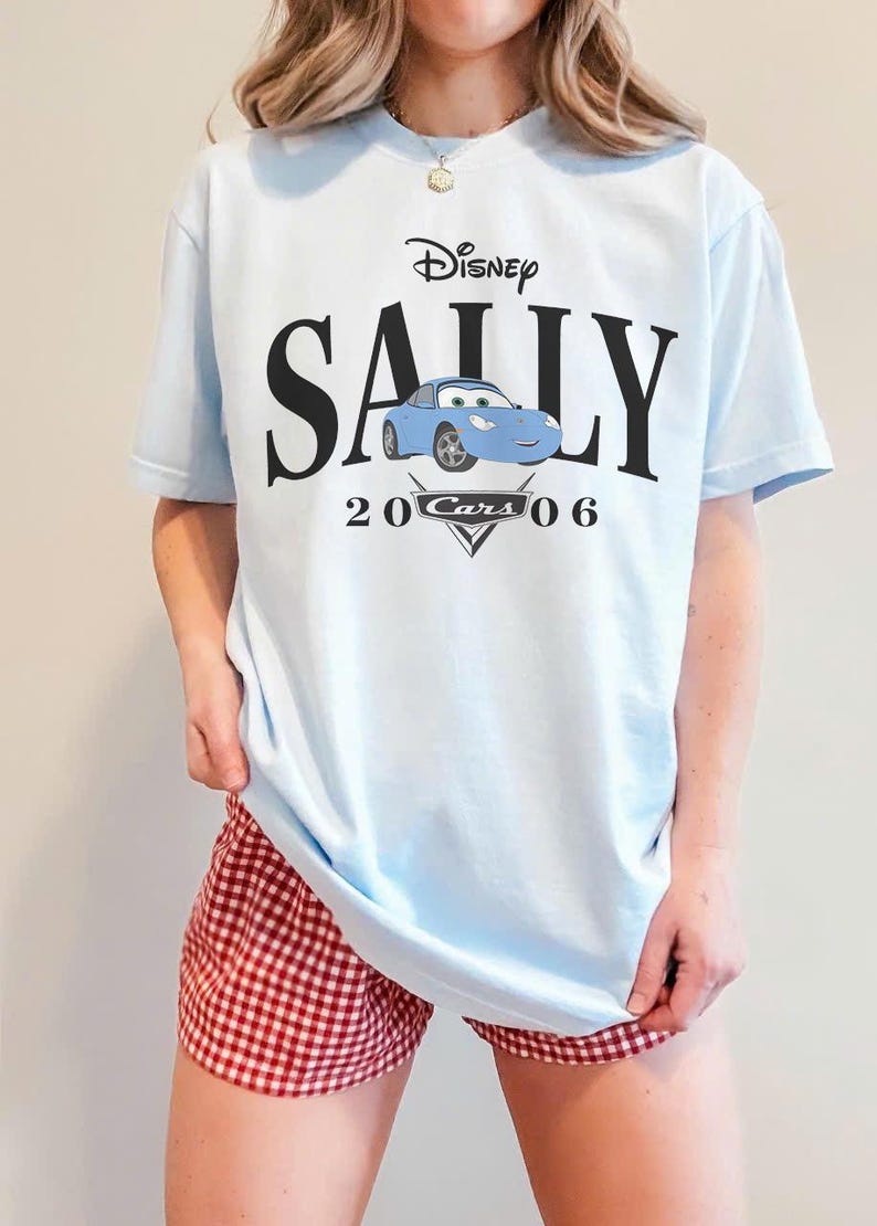 Op de afbeelding: Lichtblauw T-shirt met het Disney Cars-personage Sally. Het shirt heeft het Disney-logo boven de naam "SALLY" in grote zwarte letters, met "2006" en een autografiek eronder. De persoon draagt een rood-wit geruite short.