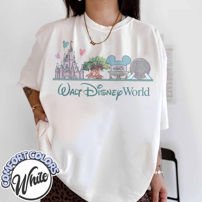 May include: White Comfort Colors t-shirt featuring a pastel graphic of Disney World landmarks, including a castle, tree, and Spaceship Earth. The words "Walt Disney World" are printed below the graphic in a teal script. The shirt has a relaxed fit.