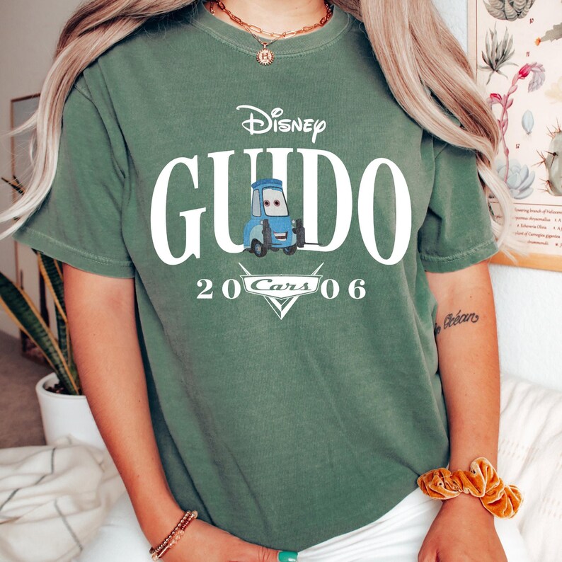 Op de afbeelding: Groen t-shirt met het Disney Cars-personage Guido. Het ontwerp bevat het Disney-logo, de naam "Guido" in het wit, een blauwe Guido-afbeelding en de tekst "20 Cars 06" in het wit.