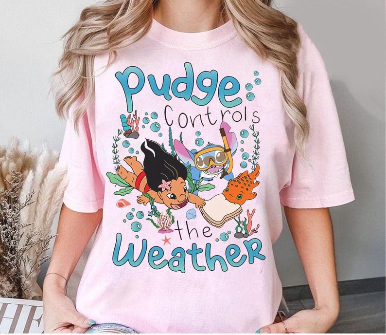 May include: A pale pink t-shirt with a cartoon illustration of Lilo and Stitch underwater, accompanied by the text "Pudge Controls the Weather" in blue. The design incorporates bubbles, coral, and a fish.