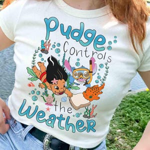 May include: A cream-coloured t-shirt with a colourful graphic featuring the text "Pudge Controls the Weather." The design includes cartoon characters and aquatic elements. A casual, everyday wear item.