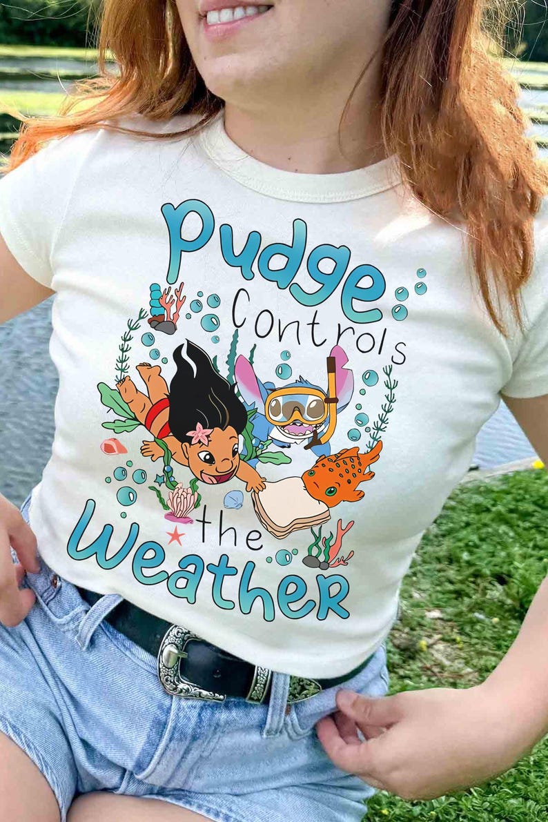 May include: A cream-coloured t-shirt with a colourful graphic featuring the text "Pudge Controls the Weather." The design includes cartoon characters and aquatic elements. A casual, everyday wear item.