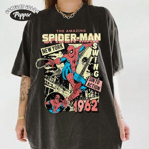 May include: Dark gray t-shirt featuring a comic book style graphic of Spider-Man swinging. The design includes the text "THE AMAZING SPIDER-MAN", "NEW YORK", "SWING", "SIN TO ACTION", and the year "1962". The shirt has a vintage, distressed look.