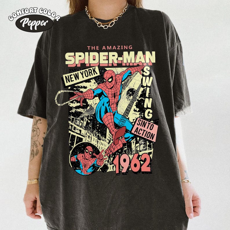 May include: Dark gray t-shirt featuring a comic book style graphic of Spider-Man swinging. The design includes the text "THE AMAZING SPIDER-MAN", "NEW YORK", "SWING", "SIN TO ACTION", and the year "1962". The shirt has a vintage, distressed look.