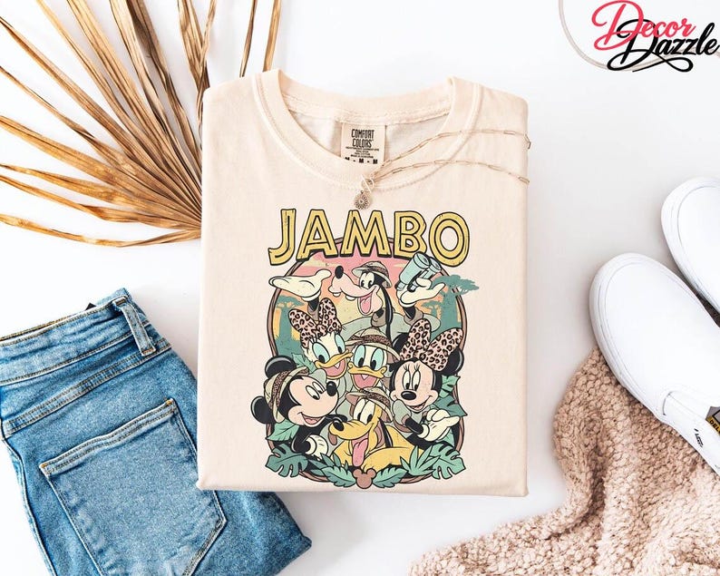 Puede incluir: Camiseta de color crema con un gr&aacute;fico colorido con la palabra "JAMBO" en letras amarillas. El gr&aacute;fico incluye personajes de dibujos animados como Mickey y Minnie Mouse, Donald y Daisy Duck y Goofy. La camiseta se combina con vaqueros azules y zapatillas blancas.