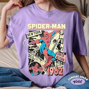 May include: A violet-colored t-shirt featuring a vintage Spider-Man graphic. The design includes the text "THE AMAZING SPIDER-MAN," "NEW YORK," and "1962." The graphic depicts Spider-Man in action with a cityscape background. The shirt is labeled "COMFORT COLOR Violet."