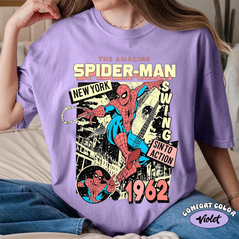 May include: A violet-colored t-shirt featuring a vintage Spider-Man graphic. The design includes the text "THE AMAZING SPIDER-MAN," "NEW YORK," and "1962." The graphic depicts Spider-Man in action with a cityscape background. The shirt is labeled "COMFORT COLOR Violet."