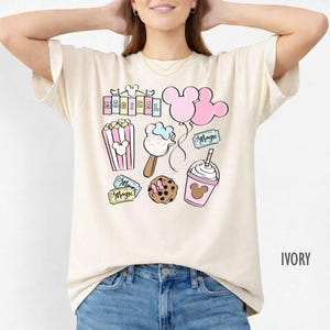 May include: Ivory-colored t-shirt featuring colorful cartoon graphics of popcorn, ice cream, balloons, a cookie, and a drink, all with a Disney theme. The word "MAGICAL" is printed at the top. The shirt is paired with blue jeans.
