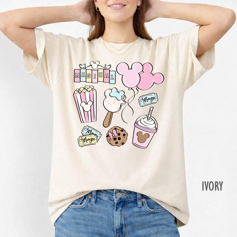 May include: Ivory-colored t-shirt featuring colorful cartoon graphics of popcorn, ice cream, balloons, a cookie, and a drink, all with a Disney theme. The word "MAGICAL" is printed at the top. The shirt is paired with blue jeans.