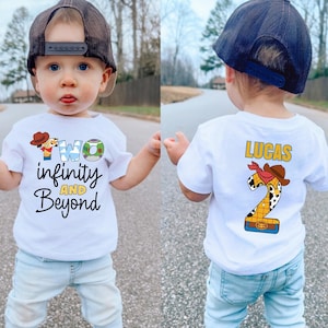 May include: A white short-sleeved t-shirt featuring a graphic print. The front displays the text "TWO infinity AND Beyond" with character-themed letters. The back shows the name "LUCAS" above a large number "2" decorated with a cowboy hat.