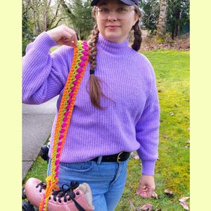 May include: A pair of pink roller skates with black wheels and laces, held by a colorful strap. The strap is crocheted with pink, orange, and green yarn. The skates are paired with a purple sweater and blue jeans.