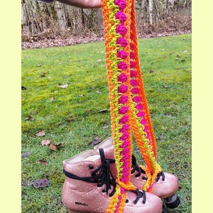 May include: Pink glitter roller skates with black laces and wheels. A vibrant, crocheted strap in orange, pink, and green is attached to the skates. The text "@knottyllamas" is visible on the skates.