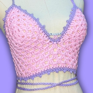 May include: A handmade pink crochet top with a lilac trim and straps. The top features the word "WITCH" spelled out in white beads. The top is tied at the waist with a lilac cord. The crochet design is open and airy.