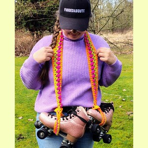 Peut inclure: Une paire de patins à roulettes roses scintillants avec des roues et des lacets noirs. Ils sont maintenus par une sangle en crochet orange, jaune et rose. Une casquette noire avec le texte "FUGGEDABOUTIT" est portée.