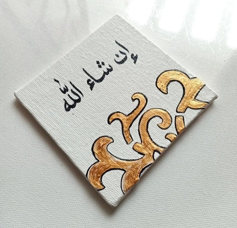 May include: A white canvas with black calligraphy that reads "In Sha Allah" and a gold decorative design in the corner.