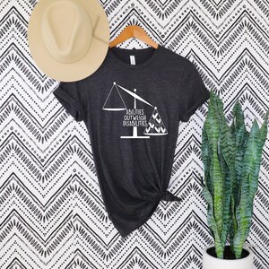 May include: Dark gray t-shirt with the text "ABILITIES OUTWEIGH DISABILITIES" in white. The design features a scale with the words on either side. A beige hat hangs above the shirt, and a potted plant is to the right.
