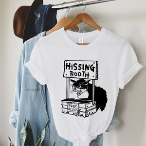 Pode incluir: Camiseta branca com uma ilustração em preto e branco de um gato a sibilar numa cabine. A placa acima diz "HISSING BOOTH" e a cabine diz "FREE HISSES". A camisa está pendurada em um cabide de madeira.