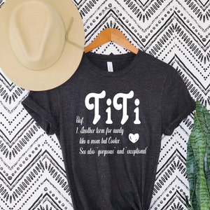 May include: Dark gray t-shirt with the word "TiTi" in white script. The definition below reads "Another term for aunty like a mom but Cooler." A tan hat hangs above the shirt.