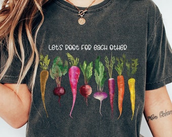 May include: Dark gray t-shirt with the text "Let's root for each other" above a watercolor illustration of various root vegetables. The vegetables include carrots, beets, and radishes in shades of orange, red, and purple.