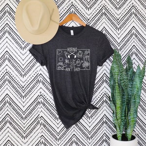 Op de afbeelding: Donkergrijs t-shirt met de witte tekst "HOEING AIN'T EASY" in een rechthoekige afbeelding. De afbeelding bevat tuingereedschap. Een beige hoed hangt erboven. Een plant in een pot staat rechts.