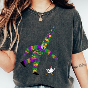 May include: Dark gray t-shirt featuring a colorful graphic of a Mardi Gras jester with a chicken. The jester is depicted in purple, green, and yellow, with a white face and black shoes. The chicken is white with red accents.