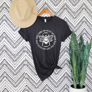 May include: Dark gray t-shirt with a white bee graphic and the words "YOURSELF - HUMBLE - KIND - POSITIVE - FUN - CONFIDENT - BRAVE - SMART - HAPPY - WILD - CREATIVE - JOYFUL" around the bee. A beige hat hangs above the shirt.