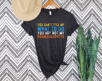 May include: Dark gray t-shirt with the text "YOU CAN'T TELL ME WHAT TO DO YOU ARE NOT MY GRANDDAUGHTER" in blue, orange, and yellow. A beige hat hangs above the shirt on a wooden hanger. The background is a black and white zigzag pattern.