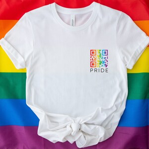 May include: White t-shirt with a rainbow QR code and the word "PRIDE" printed below it. The shirt is displayed on a rainbow flag background, symbolizing LGBTQ+ pride and inclusivity. The t-shirt is a casual wear item.