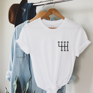 Könnte beinhalten: Weißes T-Shirt mit einem schwarzen Schalthebel-Motiv. Die Grafik zeigt den Buchstaben "R", der die Gänge darstellt. Das Shirt hängt an einem Holzbügel mit anderen Kleidungsstücken im Hintergrund.