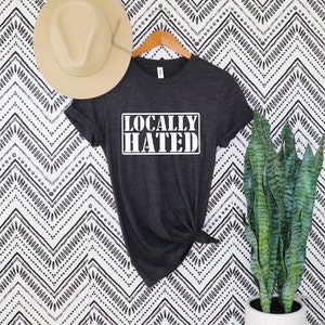 Puede incluir: Camiseta gris oscuro con el texto "LOCALLY HATED" en un rectángulo blanco. Un sombrero beige cuelga sobre la camiseta en una percha de madera. Una planta verde alta en una maceta blanca está a la derecha de la camiseta.