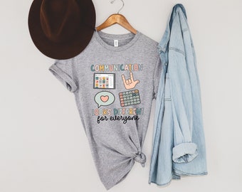 Puede incluir: Una camiseta gris jaspeado con el texto "COMMUNICATION LOOKS DIFFERENT for everyone". La camiseta presenta gráficos coloridos de una tableta, una mano haciendo el signo "Te amo", una calculadora y un bocadillo con un corazón.