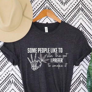 May include: Dark grey t-shirt with white text and a skeleton hand holding a cannabis leaf. The text reads "SOME PEOPLE LIKE TO stir the pot I PREFER to smoke it."