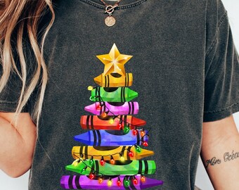 Könnte beinhalten: Dunkelgraues T-Shirt mit einem Weihnachtsbaum-Design aus bunten Buntstiften. Der Baum ist mit einem goldenen Stern gekrönt und mit Lichterketten geschmückt. Ein festliches und kreatives Design für die Weihnachtszeit.