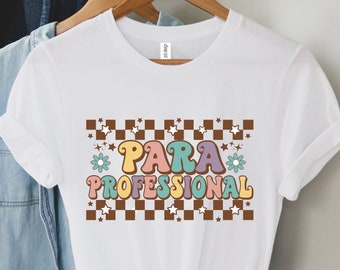 May include: White t-shirt featuring the text "PARA PROFESSIONAL" in a retro style. The design incorporates a brown and white chequered pattern, stars, and pastel-coloured flowers. The shirt is displayed on a wooden hanger.