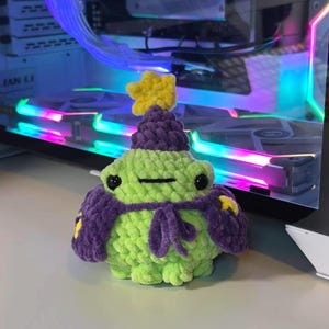 May include: A crochet frog plushie featuring a light green body, black bead eyes, and a small horizontal mouth. The frog wears a dark purple pointed hat topped with a yellow star and a matching dark purple cape tied at the front with yellow stars.