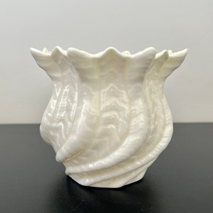 Puede incluir: Un jarrón de cerámica blanco con un diseño de concha ondulado. El jarrón está hecho de un material liso y brillante y tiene un aspecto delicado y femenino.