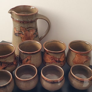 May include: A set of brown and green ceramic mugs and a jug with a brown and orange design. The mugs and jug have a rustic, handmade look.